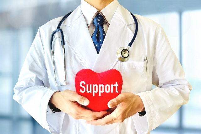 聴診器を首にかけ白衣を着た男性医師がサポートと書いたハートの形を両手で持っているところ、清潔感がある印象